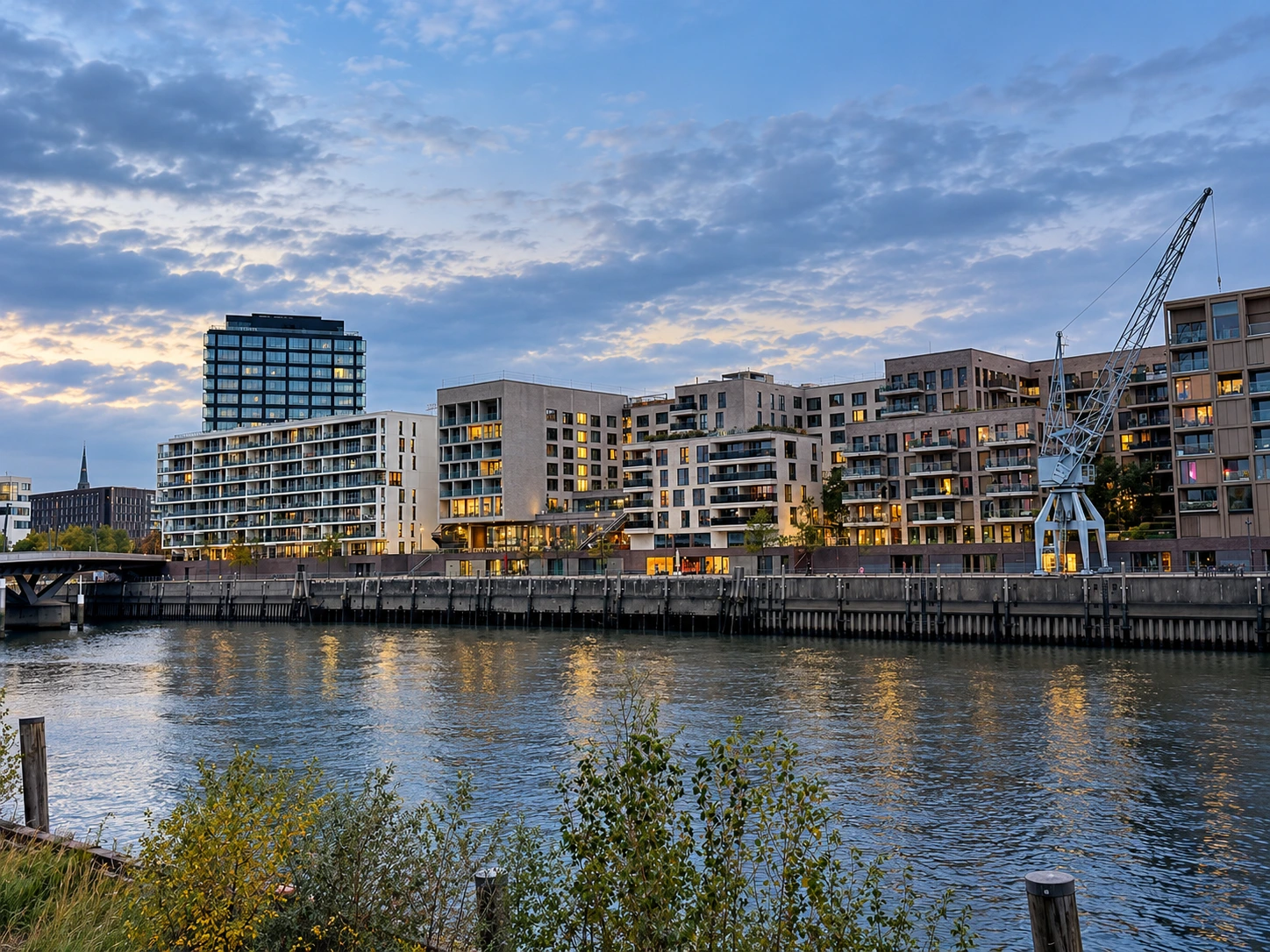 Moderne Wohngebäude am Wasser in der Hamburger HafenCity mit Hafenkran und beleuchteten Fenstern in der Abenddämmerung.